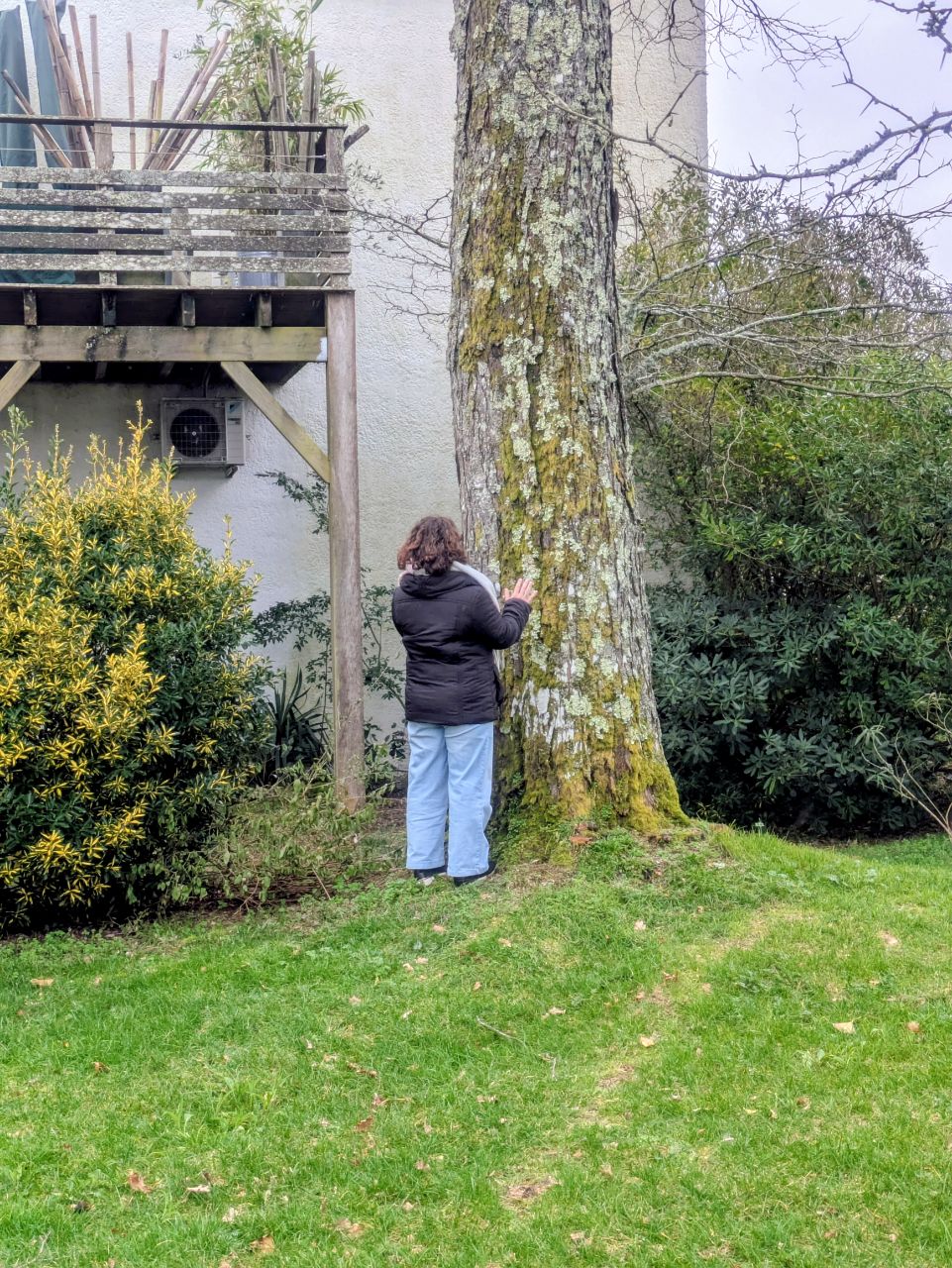 une jeune femme de dos entre en contact avec un arbre, dans le jardin d'une résidence. elle a les mains tendues vers l'écorce et est habillée d'un manteau noir, d'une écharpe beige et d'un jean bleu clair.